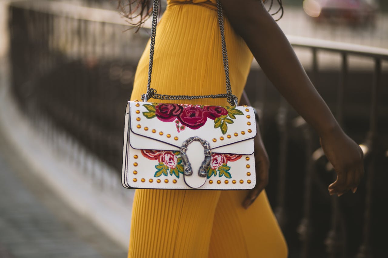 A woman in a yellow dress showcases a floral embroidered bag on a sunny day outdoors.